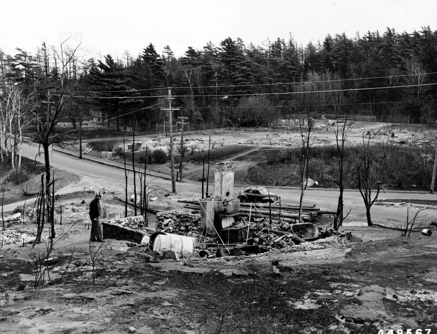 A man standing beside the chimney and burned debris of a house on Eagle Lake Road, with the road, bare lots and a stand of pines behind