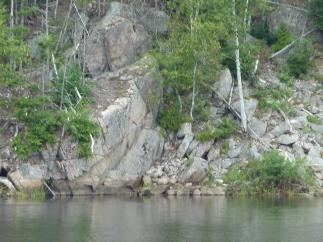 Seen from the lake, a granite cliff and talus slope drop to the water, with birch and spruce above