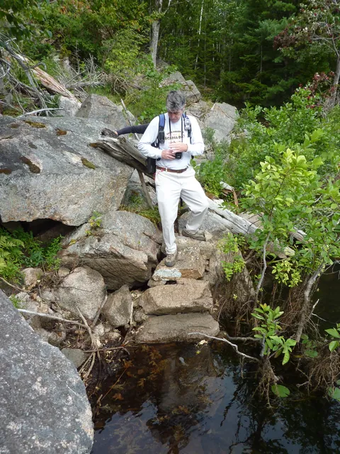 A hiker in a white shirt climbs down rough stone steps toward the water between boulders