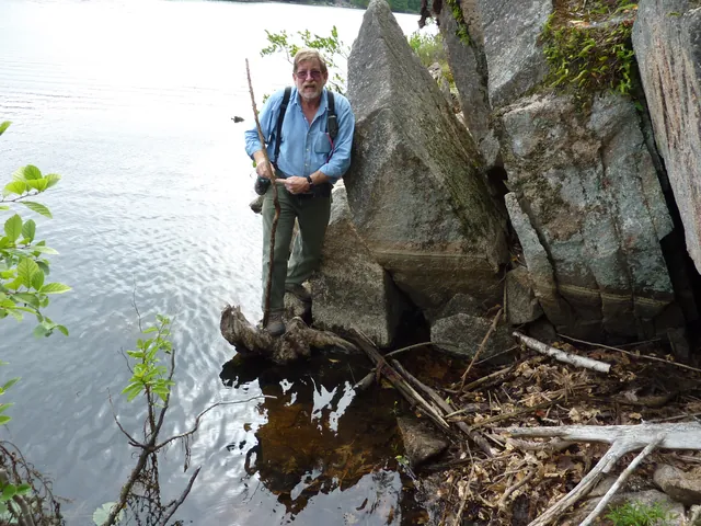 A hiker with a walking stick stands on a stone at the water's edge beside tall rocks