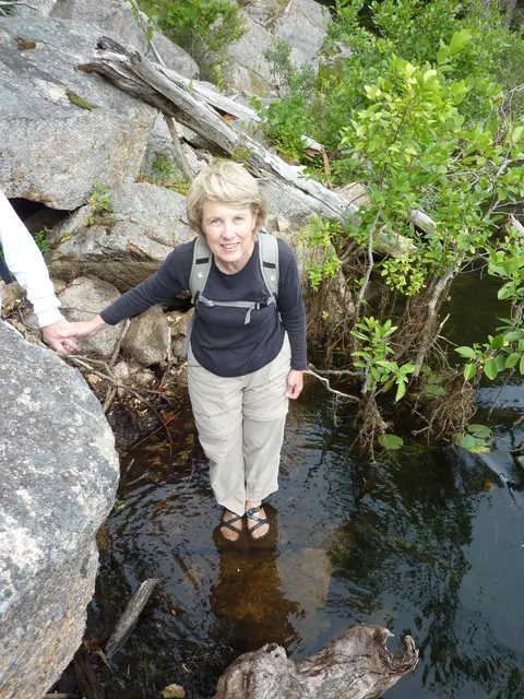 A smiling hiker stands in shallow water on the stones between boulders and brush