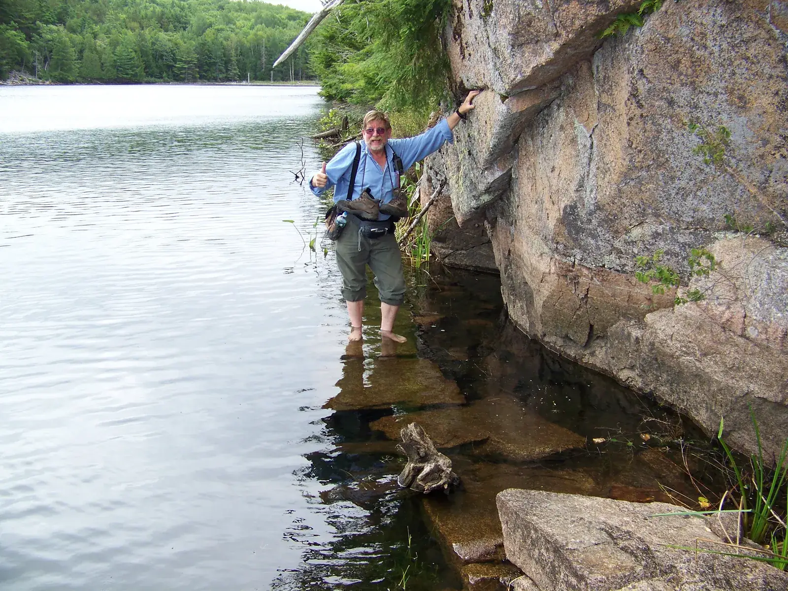 A hiker stands knee deep in clear water on submerged flat stones, steadying himself with one hand on a tall granite cliff