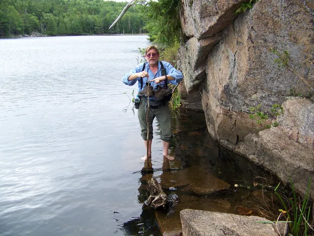 A hiker stands on a submerged stone in shallow water against the granite cliff, holding a camera