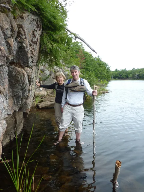Two hikers stand together on submerged stones at the cliff foot, one with a walking pole