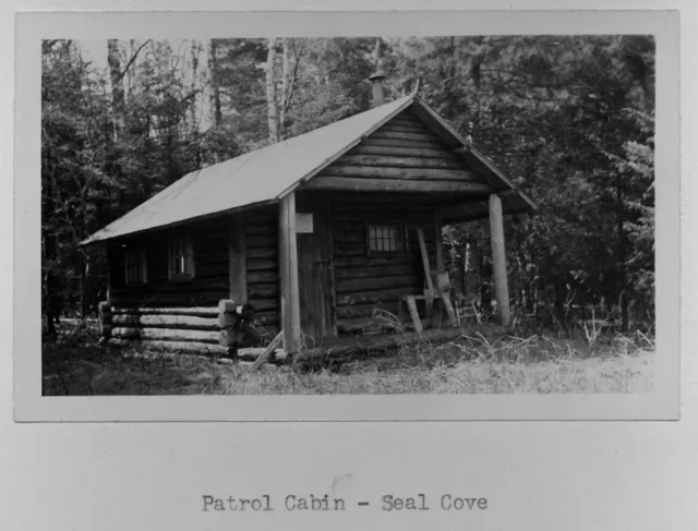 Archival print of a small log cabin with a metal roof and a covered porch, set against tall woods, captioned Patrol Cabin - Seal Cove