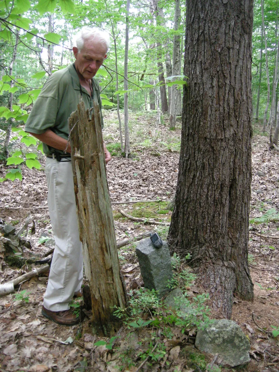 A white-haired crew member beside a broken stump and a small granite boundary stone with a GPS unit on it