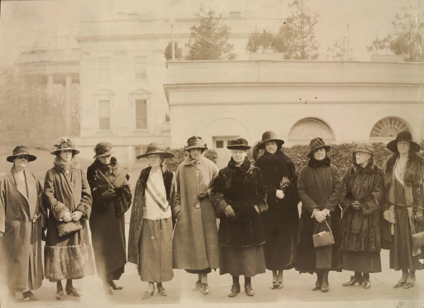 Ten women in hats and fur-trimmed coats standing in a row outside the White House