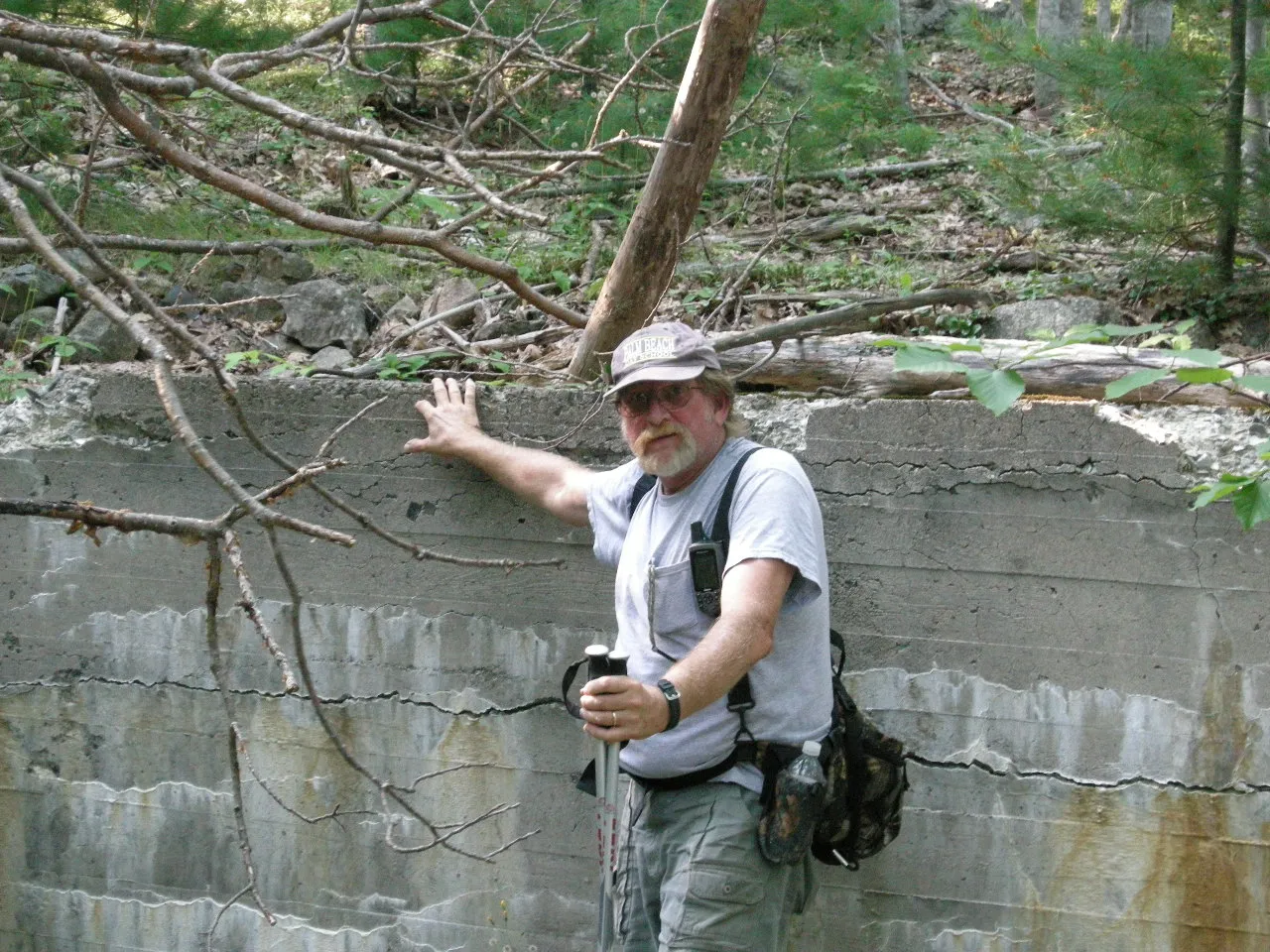 A crew member in a cap stands beside a tall concrete foundation wall in the woods